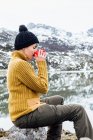 Side view tranquil young female in warm sweater and hat drinking hot beverage while sitting on sharp stone on cold lake shore surrounded by rough severe snowy mountains — Stock Photo