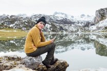Side view full body young glad female wearing warm sweater and hat using mobile phone and looking away while sitting on sharp stony lake shore surrounded by severe snowy mountains — Stock Photo