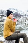 Вид сбоку спокойная молодая женщина в теплом свитере и шляпе пьет горячий напиток, сидя на резком камне на холодном берегу озера в окружении суровых суровых снежных гор — стоковое фото