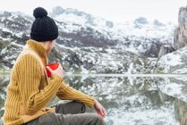 Back view of anonymous tranquil young female in warm sweater and hat drinking hot beverage while sitting on sharp stone on cold lake shore surrounded by rough severe snowy mountains — Stock Photo