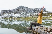 Женщина в тёплом свитере и черной шляпе сидит с вытянутыми руками на грубом камне на спокойном озере с закрытыми глазами рядом с пейзажем снежных скалистых гор — стоковое фото