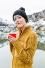 Retrato de jovem mulher tranquila em suéter quente e chapéu segurando bebida quente na frente de montanhas nevadas — Fotografia de Stock