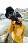 Жінка-фотограф в теплому одязі і капелюсі знімає фотографії і дивиться на камеру, стоячи на березі озера в оточенні грубих засніжених гір — стокове фото