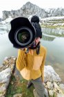 Жінка-фотограф в теплому одязі і капелюсі знімає фотографії і дивиться на камеру, стоячи на березі озера в оточенні грубих засніжених гір — стокове фото