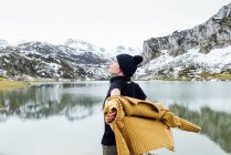 Visão traseira atraente alegre fêmea em suéter quente e chapéu estendendo os braços em emoção e olhando para longe, enquanto em pé na costa do lago ondulando frio cercado por montanhas nevadas rochosas — Fotografia de Stock