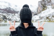 Anonymous female hiding face behind black hat and showing v sign while standing on snowy rough highlands on lake shore — Stock Photo