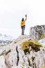 Volver ver fotógrafo de cuerpo completo en ropa de abrigo levantando el brazo con cámara fotográfica y de pie sobre roca rugosa rígida en las tierras altas nevadas de Asturias - foto de stock