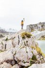 Просмотр полного тела фотографа в теплой одежде, поднимающего руку с фотокамерой и стоящего на жесткой грубой скале в снежных горах в Астурии — стоковое фото