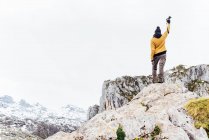 Просмотр полного тела фотографа в теплой одежде, поднимающего руку с фотокамерой и стоящего на жесткой грубой скале в снежных горах в Астурии — стоковое фото