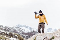 Ganzkörperfotografin in warmer Kleidung hebt Arm mit Fotokamera und steht auf steifem, rauem Felsen im verschneiten Hochland in Asturien — Stockfoto