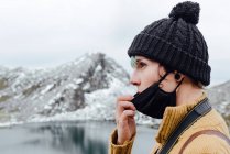 Side view serene young female traveler in warm hat lowering face mask and looking away in contemplation while standing on cold lake coast against majestic snowy mountain ridge — Stock Photo