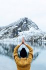 Невпізнавана жінка в теплому одязі і капелюсі, що медитує на Лотос Посе з руками Намасте на озері проти суворих снігових гір в Астурії. — стокове фото