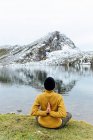 Rückansicht Unerkennbare Frau in warmer Kleidung und Hut meditiert auf Lotus Pose mit Namaste Händen auf dem Rücken am Seeufer vor schwer verschneiten Bergen in Asturien — Stockfoto