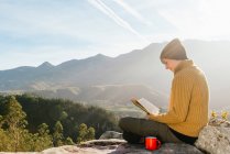 Seitenansicht des Inhalts weibliche Reisende sitzt mit einer Tasse Heißgetränk und liest interessante Buch vor dem Hintergrund der spektakulären Berglandschaft an sonnigen Tag — Stockfoto