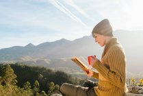 Vista lateral del contenido viajero femenino sentado con una taza de bebida caliente y leyendo un interesante libro sobre el fondo del espectacular paisaje montañoso en un día soleado - foto de stock