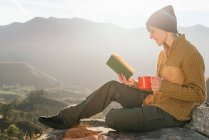 Vue latérale du contenu voyageur féminin assis avec une tasse de boisson chaude et la lecture livre intéressant sur fond de paysage montagneux spectaculaire par jour ensoleillé — Photo de stock