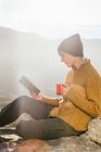 Боковой вид женщины, сидящей за чашкой горячего напитка и читающей интересную книгу на фоне живописного горного пейзажа в солнечный день — стоковое фото