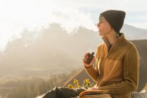 Vista lateral do viajante feminino tranquilo sentado na rocha enquanto fuma vape e exala nuvem de fumaça durante as férias nas montanhas — Fotografia de Stock