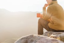 Rückansicht des Wanderers, der auf einem Stein sitzt und an einem sonnigen Tag die atemberaubende Landschaft des Hochlandtals beobachtet, während er Kaffee trinkt — Stockfoto