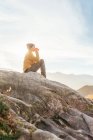 Vista lateral del excursionista sentado en piedra y observando increíbles paisajes del valle de las tierras altas en un día soleado mientras bebe taza de café - foto de stock