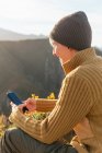 Боковой вид позитивной женщины, сидящей на мобильном телефоне на фоне горного хребта в солнечный день — стоковое фото