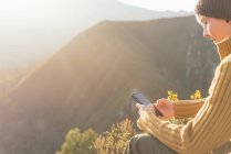 Side view of positive female explorer messaging on mobile phone while sitting on background of mountain range on sunny day — Stock Photo