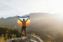 Vista trasera del excursionista de pie con la bandera LGBT arco iris con inscripción Paz y disfrutar de la libertad con los brazos extendidos en las montañas - foto de stock