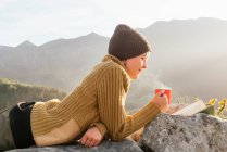 Vista lateral del contenido viajero femenino acostado con una taza de bebida caliente y leyendo un interesante libro sobre el fondo del espectacular paisaje montañoso en un día soleado - foto de stock