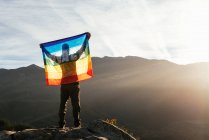 Vista trasera del excursionista de pie con la bandera LGBT arco iris con inscripción Paz y disfrutar de la libertad con los brazos extendidos en las montañas - foto de stock