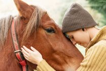 Side view of tender female stroking muzzle of chestnut horse grazing in meadow while spending weekend in countryside — Stock Photo