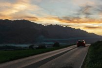 Автомобильная прогулка по асфальтированной дороге на фоне горного хребта под закатным небом вечером — стоковое фото