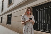 Unrecognizable female tourist in dress and mask text messaging on cellphone on pavement near stone building in Seville — Stock Photo
