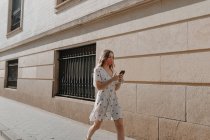Unerkennbare Touristin in Kleid und Maske mit SMS auf Handy auf Bürgersteig in der Nähe eines Steingebäudes in Sevilla — Stockfoto