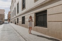 Неузнаваемая женщина-туристка в одежде и маске текстовых сообщений на мобильном телефоне на тротуаре возле каменного здания в Севилье — стоковое фото