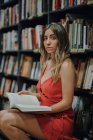 Вид збоку молодої жінки в червоному вечірньому одязі з відкритим підручником, сидячи в книгарні і дивлячись на камеру — стокове фото