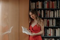 Vista laterale della giovane donna in prendisole rosso con libro di testo aperto in piedi in libreria e guardando la fotocamera — Foto stock