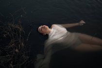 De cima jovem morena serena em calcinha molhada e pano branco translúcido deitado com os olhos fechados em água tranquila lagoa escura — Fotografia de Stock