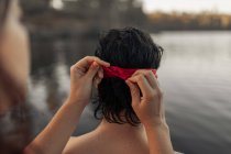 Cultivo anónimo mujer turista atando la venda roja en la cabeza de la pareja contra el agua ondulada durante el viaje - foto de stock