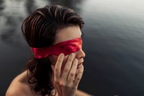Vista lateral de turista femenina gentil anónima con los ojos vendados rojos tocando la cara contra el agua a la luz del día - foto de stock