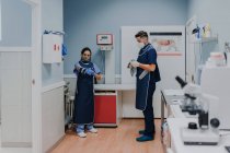 Veterinario masculino joven irreconocible con máscara estéril y guantes de pie cerca de una colega femenina mientras se prepara para el trabajo en el laboratorio - foto de stock