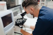 Side view of crop anonymous male medic in uniform and mask using microscope while working in lab — Stock Photo