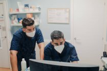 Jovens médicos anônimos em máscaras respiratórias e uniformes trabalhando no computador desktop na clínica — Fotografia de Stock