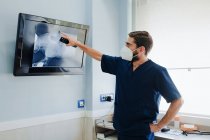 Veterinario masculino anónimo en máscara respiratoria y uniforme explicando anatomía del animal mamífero mientras toca la pantalla con ilustración de rayos X en clínica - foto de stock