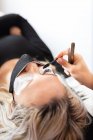 Esthéticienne professionnelle appliquant des cils artificiels sur une jeune cliente portant un masque protecteur dans un studio de beauté moderne — Photo de stock