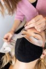 Профессиональная женщина-мастер с пинцетом наносит искусственные ресницы на молодую клиентку в маске для лица в светлом современном салоне — стоковое фото