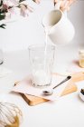 Cultivo persona anónima verter leche tibia fresca de la jarra en el vidrio colocado en la tabla de cortar en la cocina ligera - foto de stock