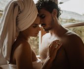 Tender woman with towel wrapped around head embracing gently bearded boyfriend with closed eyes near window with water drops in shack — Stock Photo