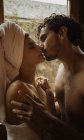 Femme tendre avec serviette enroulée autour de la tête embrassant petit ami doucement barbu avec les yeux fermés près de la fenêtre avec des gouttes d'eau dans la cabane — Photo de stock