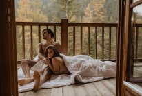 Romantic undressed couple lying on cozy blanket and cuddling on wooden cottage terrace against deciduous forest on autumn — Stock Photo