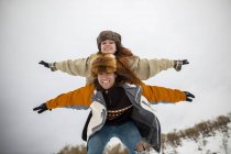 Allegro turista maschio che trasporta la ragazza con le braccia sollevate a cavalluccio sul monte innevato in inverno — Foto stock